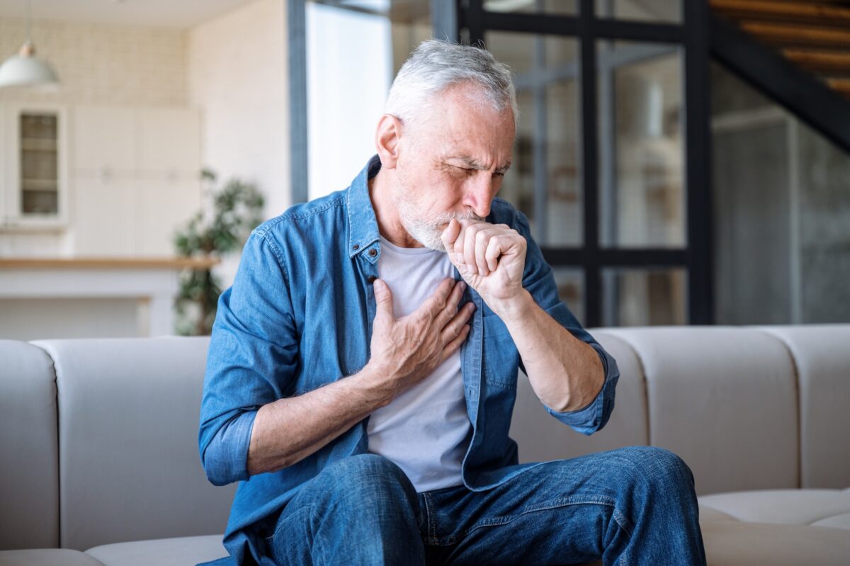 Senior man coughing while sitting on sofa at home and holding hand on chest.